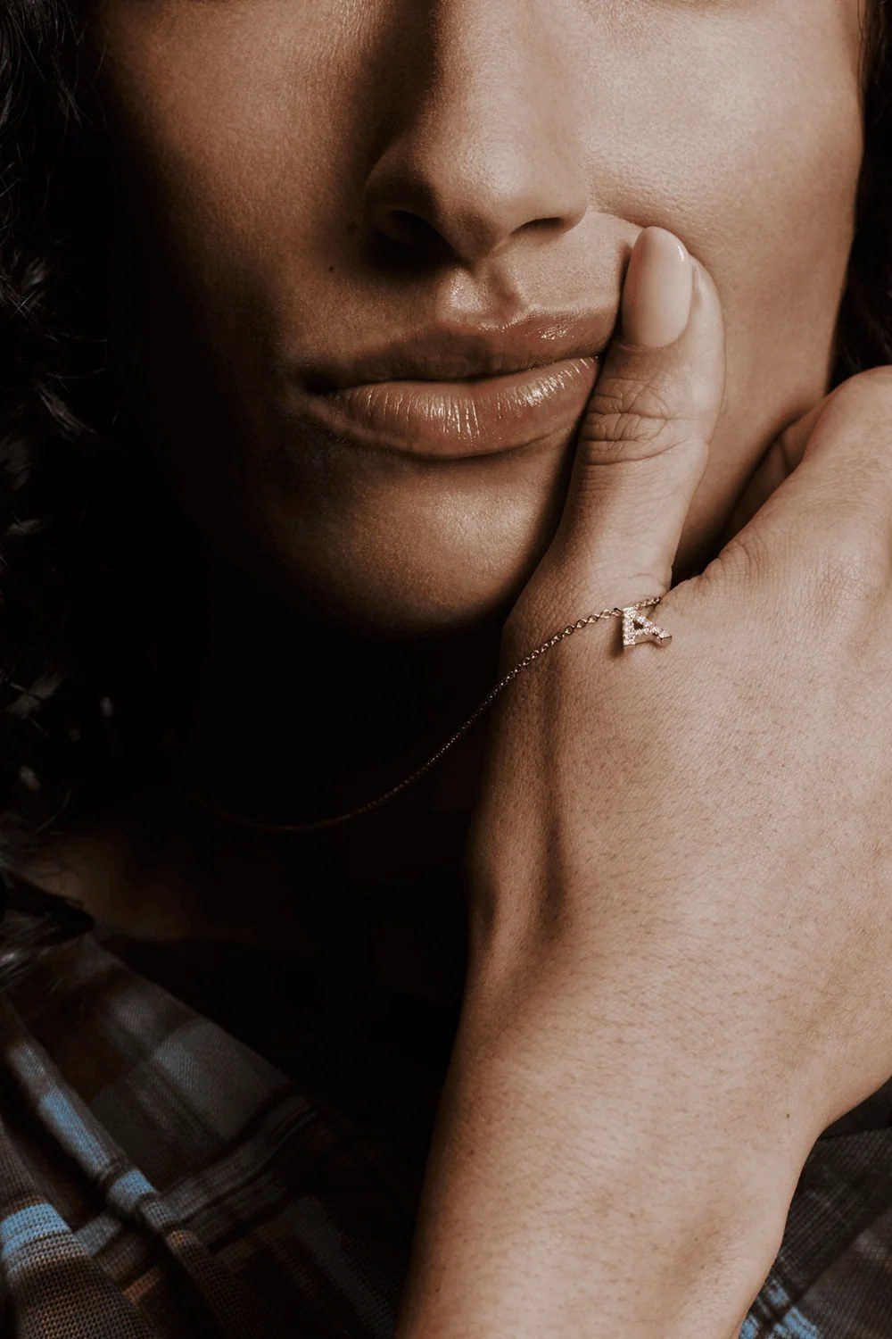 A hand resting against a face, wearing a pavé initial chain bracelet in gold
